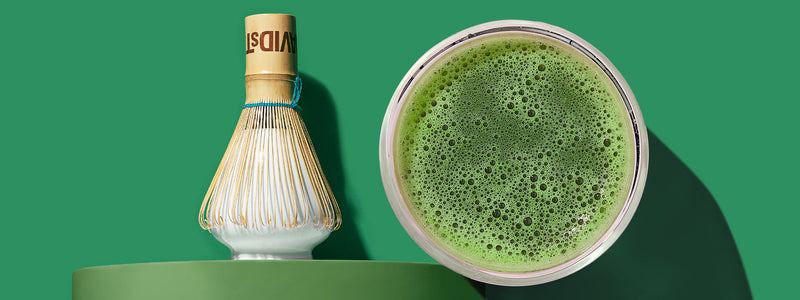 A bird's-eye view of frothy green matcha next to a bamboo whisk on a whisk holder against a dark green background.
